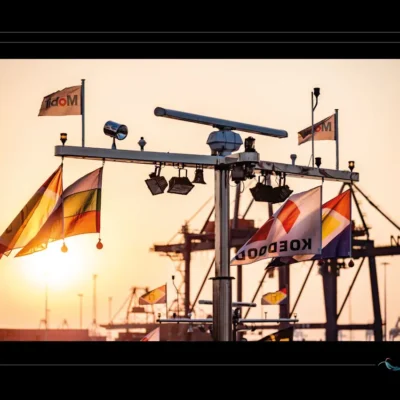 Photo poétique de port au coucher du soleil – pavillons maritimes et infrastructures industrielles. Explorez l’esthétique du transport maritime à travers une image vibrante mêlant mâts, drapeaux, grues et lumière dorée. Idéal pour illustrer les ports bretons, la logistique navale et la beauté des instants suspendus entre mer et industrie. Ordigraph