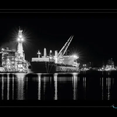 Photo artistique noir et blanc d’un port industriel la nuit – cargo illuminé et reflets maritimes. Explorez l’atmosphère poétique d’un terminal portuaire en pleine obscurité, où la lumière sculpte les formes et le silence révèle la grandeur. Idéal pour illustrer les métiers de la mer, la logistique nocturne et l’esthétique industrielle. Ordigraph
