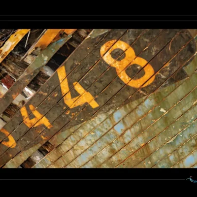Photo artistique d’un bateau ancien, bois en diagonale, peinture jaune-orange, immatriculation 448 effacée. Texture marine, contraste visuel fort. Idéal pour blog sur patrimoine nautique, design vintage ou inspiration artistique. Mots-clés : bateau abandonné, numéro 448, bois vieilli, peinture diagonale, photographie maritime, textures marines, ordigraph