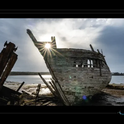 Photo artistique d’une épave marine sur vase, bois ouvert, lumière traversante, ciel nuageux, ambiance portuaire abandonnée. Idéal pour blog sur patrimoine maritime, design nostalgique ou inspiration artistique. Mots-clés : épave bateau, bois éventré, lumière traversante, vase portuaire, photographie artistique, textures maritimes, Ordigraph