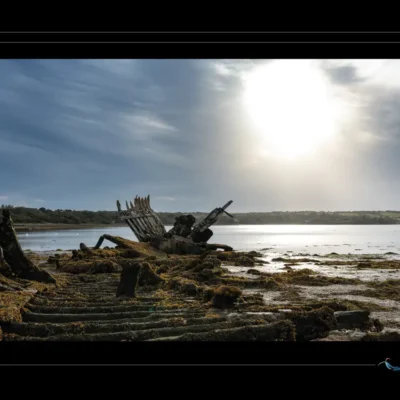 Photo artistique d’une épave marine rongée par le temps, bois et métal dégradés, algues et mousse, lumière dramatique. Idéal pour blog sur patrimoine maritime, design nostalgique ou inspiration artistique. Mots-clés : épave bateau, bois rongé, algues marines, lumière dramatique, photographie artistique, textures portuaires, Ordigraph
