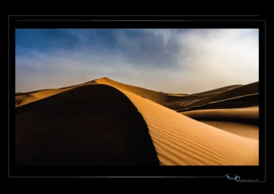 Photographie artistique de désert — crête de dune, contraste lumière/ombre, textures naturelles, ciel nuageux. Image fine art signée Ordigraph, idéale pour décoration murale, galerie photo, blog de voyage ou contenu éditorial sur les paysages extrêmes et la beauté minérale du désert.