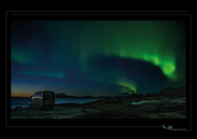 Photo nocturne d’une aurore boréale au-dessus d’un paysage nordique. Le ciel est traversé par des traînées vertes intenses, dynamiques et vibrantes, qui s’élèvent au-dessus d’un plan d’eau calme bordé de roches. Un van est stationné sur la droite, ajoutant une présence humaine discrète face à l’immensité. Les étoiles scintillent, et les montagnes à l’horizon se découpent sous la lumière céleste. La photo joue sur la tension entre solitude terrestre et éclat cosmique, entre immobilité et jaillissement. Une scène où le ciel semble parler. Ordigraph