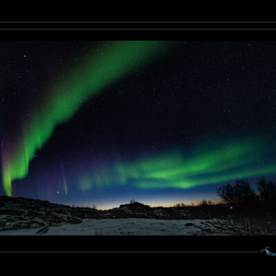 Photo nocturne d’une aurore boréale au-dessus d’un paysage arctique enneigé. Le ciel est parcouru de traînées vertes et violettes, fluides et vibrantes, qui s’élèvent comme des fibres lumineuses issues du souffle solaire. Le sol est composé de neige, de roches et de végétation rare, silhouetté contre la lumière céleste. Les étoiles scintillent dans un ciel limpide, renforçant l’ambiance cosmique. La photo joue sur la tension entre matière terrestre et mouvement céleste, entre immobilité et fluidité. Une scène où la lumière devient textile céleste. - Ordigraph