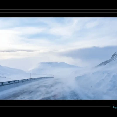 Route de montagne enneigée vers le Cap Nord — vent fort, neige soufflée, poteaux rouges, rambarde, ciel nuageux. Photo de paysage norvégien extrême, ambiance dramatique et hivernale. Idéal pour galerie WordPress sur routes arctiques et navigation en milieu polaire. Ordigraph