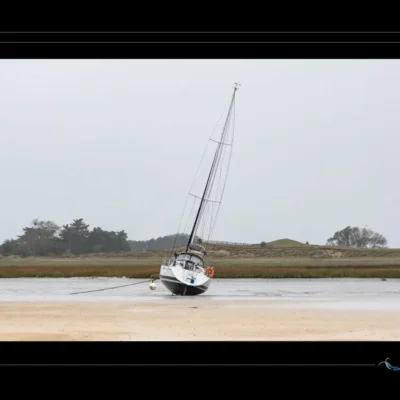 Voilier retenu par chaîne sur plage – photo poétique, bateau incliné, vent, marée basse, lumière douce. Image idéale pour galerie maritime, Bretagne, tension entre départ et attache. Voilier sur sable, chaîne d’amarrage, vent, photographie contemplative, aventure empêchée. Ordigraph