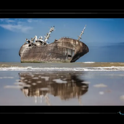 Épave du navire Que Sera Sera échouée sur une plage marocaine, avec reflet visible dans le sable humide. Histoire du navire : origine islandaise, impact économique à Laâyoune, disparition de l’équipage, rôle dans les migrations clandestines. Photographie artistique mettant en valeur la corrosion, le reflet, et la charge symbolique du lieu. Ordigraph