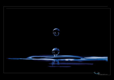 Photo à haute vitesse capturant l’instant rare où deux gouttes d’eau se croisent : l’une en chute libre, l’autre en rebond. La surface liquide révèle des ondulations fines et un éclat discret. L’éclairage bleuté sur fond noir accentue la transparence et la tension superficielle, offrant une composition verticale et vibrante. Ce cliché illustre la dynamique fluide, la gravité et la mémoire du mouvement. Ordigraph