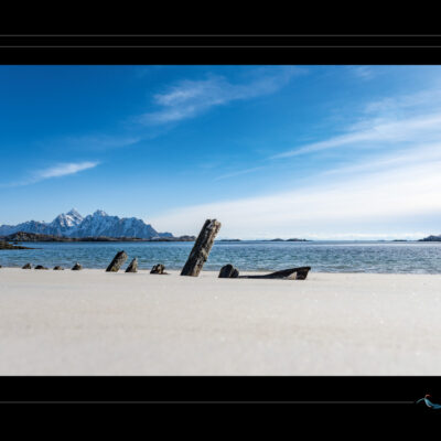 Photo de Laponie avec plage nordique, bois échoué, fjord calme, montagnes enneigées et ciel bleu. Paysage scandinave hivernal, nature sauvage, ambiance poétique et minimaliste. Idéal pour blog de voyage, site photo, inspiration nordique et récits visuels. Ordigraph