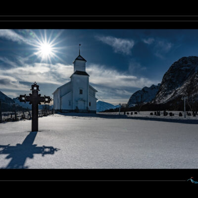 Photo hivernale de Laponie avec église blanche, croix en bois, cimetière enneigé, montagnes en arrière-plan et lumière d’hiver. Paysage nordique spirituel, ambiance calme et poétique, contraste entre ombre et neige. Idéal pour blog de voyage, site photo, inspiration scandinave et récits contemplatifs. Ordigraph