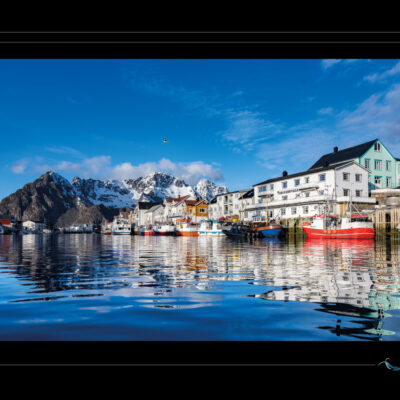 Photo du village de Henningsvær en Laponie norvégienne, port nordique avec bateaux colorés, maisons traditionnelles, montagnes enneigées et ciel bleu. Paysage scandinave haute définition, ambiance hivernale, nature calme et architecture typique. Idéal pour blog de voyage, site photo, inspiration nordique et récits visuels. Ordigraph