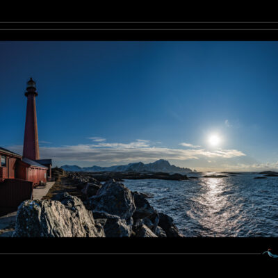 Photo du phare d’Andenes en Norvège, paysage côtier avec mer de Norvège, rochers, soleil bas, montagnes enneigées et ciel clair. Image scandinave haute définition, ambiance arctique, nature sauvage et architecture maritime. Idéal pour blog de voyage, site photo, inspiration nordique et récits visuels. Ordigraph