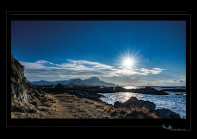 Photo de paysage côtier en Laponie avec rochers, mer de Norvège, soleil bas, montagnes enneigées et ciel clair. Image scandinave haute définition, ambiance hivernale, nature sauvage et lumière arctique. Idéal pour blog de voyage, site photo, inspiration nordique et récits poétiques. Ordigraph