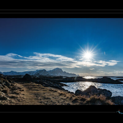 Photo de paysage côtier en Laponie avec rochers, mer de Norvège, soleil bas, montagnes enneigées et ciel clair. Image scandinave haute définition, ambiance hivernale, nature sauvage et lumière arctique. Idéal pour blog de voyage, site photo, inspiration nordique et récits poétiques. Ordigraph