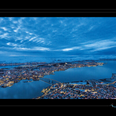 Photo de Tromsø en Norvège à l’heure bleue, vue aérienne avec fjord, pont, lumières urbaines, montagnes enneigées et ciel nuageux. Paysage scandinave haute définition, ambiance hivernale, contraste entre ville et nature. Idéal pour blog de voyage, site photo, inspiration nordique et récits visuels. Ordigraph