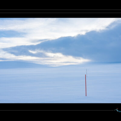 Photo hivernale en Laponie avec paysage enneigé, poteaux rouges alignés, ciel nuageux et ambiance minimaliste. Image scandinave haute définition, nature arctique, contraste visuel entre neige et signalisation. Idéal pour blog de voyage, site photo, inspiration nordique et récits contemplatifs. Ordigraph