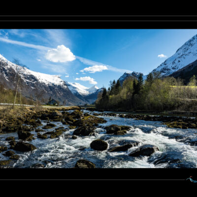 Photo de paysage norvégien avec rivière rapide, rochers, route, maison isolée, montagnes enneigées et ciel clair. Image haute définition, nature scandinave, contraste entre mouvement et calme, idéale pour blog de voyage, site photo, inspiration nordique et récits contemplatifs. Ordigraph