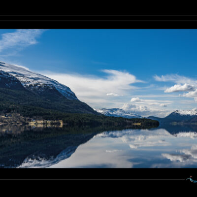 Photo de paysage nordique avec lac calme, montagnes enneigées, forêts, maisons isolées et reflets parfaits. Image haute définition, nature scandinave, symétrie naturelle, ambiance silencieuse et contemplative. Idéal pour blog de voyage, site photo, inspiration nordique et récits visuels. Ordigraph