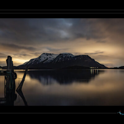 Photo nocturne de paysage nordique avec montagne enneigée, lac calme, quai en bois, ciel nuageux et lumière diffuse. Image haute définition, ambiance silencieuse et contemplative, idéale pour blog de voyage, site photo, inspiration scandinave et récits visuels. Ordigraph