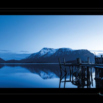 Photo de paysage nordique avec vieux quai en bois, lac calme, montagnes enneigées, ciel doux et reflets parfaits. Image haute définition, ambiance contemplative et matinale, idéale pour blog de voyage, site photo, inspiration scandinave et récits visuels. Ordigraph