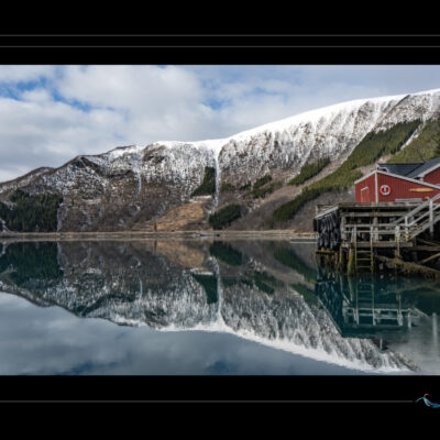 Photo de paysage nordique avec cabane rouge sur pilotis, lac calme, montagnes enneigées, ciel nuageux et reflets parfaits. Image haute définition, ambiance contemplative et isolée, idéale pour blog de voyage, site photo, inspiration scandinave et récits visuels. Ordigraph