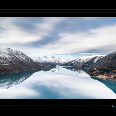 Photo de paysage nordique avec lac calme, montagnes enneigées, ciel nuageux et reflets parfaits. Image haute définition, ambiance contemplative et hivernale, idéale pour blog de voyage, site photo, inspiration scandinave et récits visuels. Ordigraph